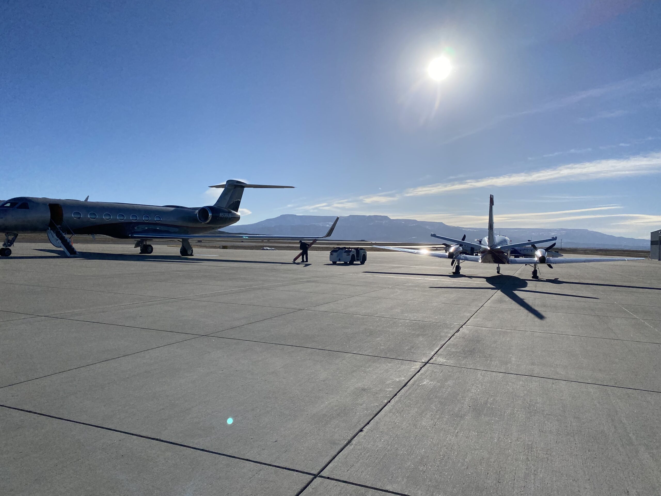 Private jet and small aircraft parked on a runway with mountains and sun
